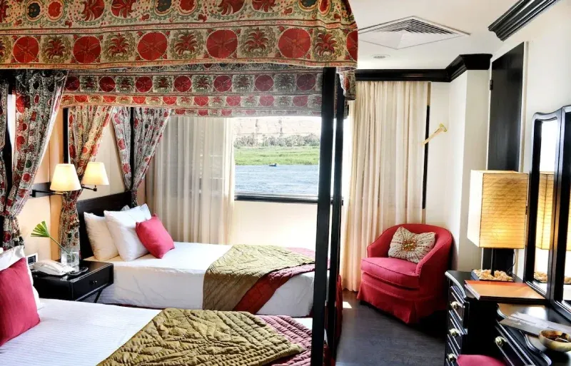 Sanctuary Sun Boat III standard cabin — four-poster bed with canopy and Egyptian linens, shuttered Nile-view windows, bathtub and shower, 1920s design, Egypt 2026